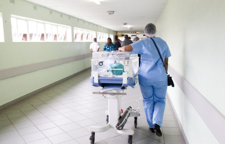A woman wearing blue scrubs walk down a hallway with a medical crib for newborns