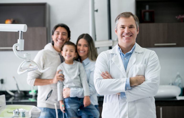 doctor-standing-and-smiling-with-arms-crossed-and-family-of-three-in-background
